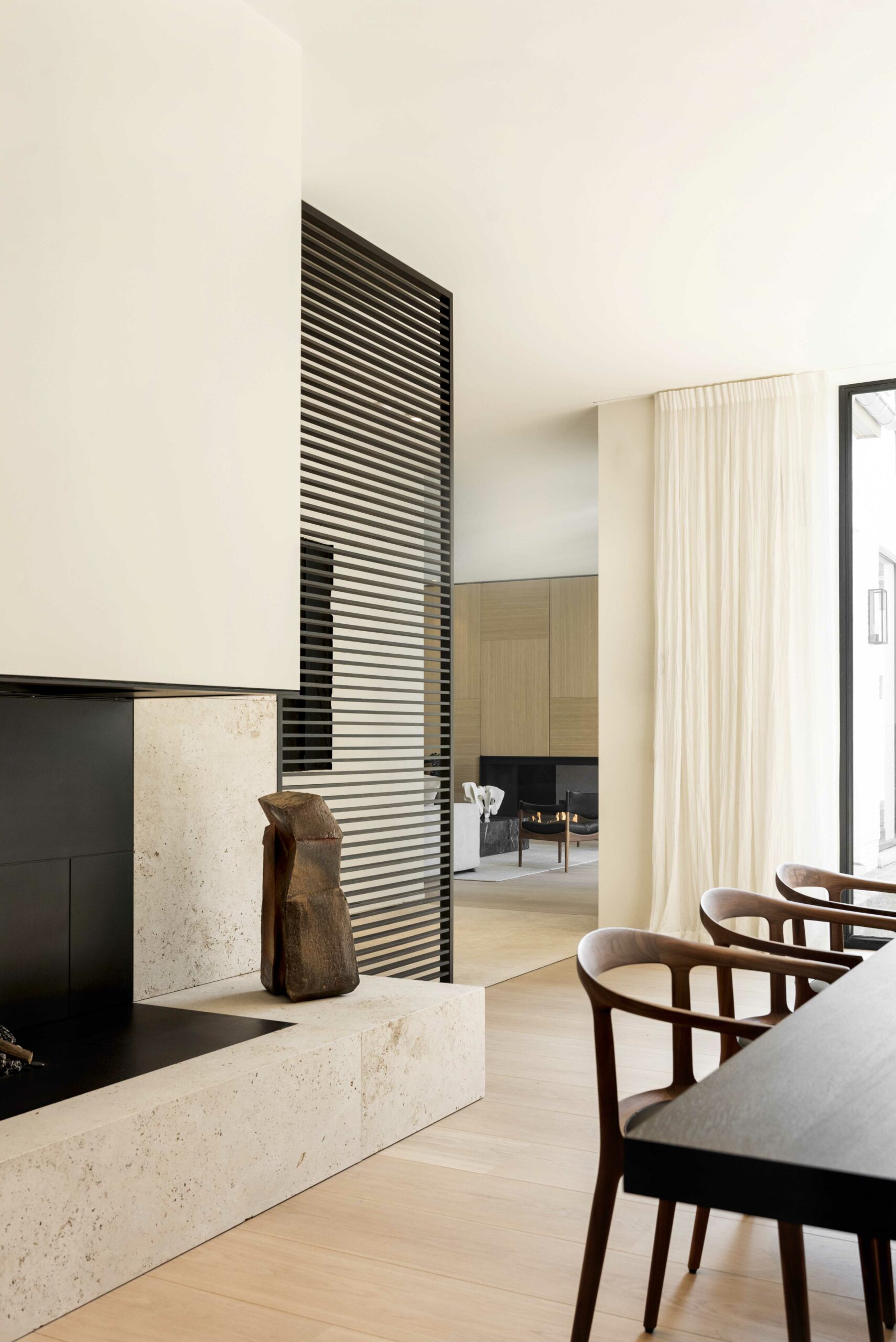 A sleek living space with a striped partition, rustic sculpture, and fireplace. Modern design meets warmth with wooden elements and sheer curtains.