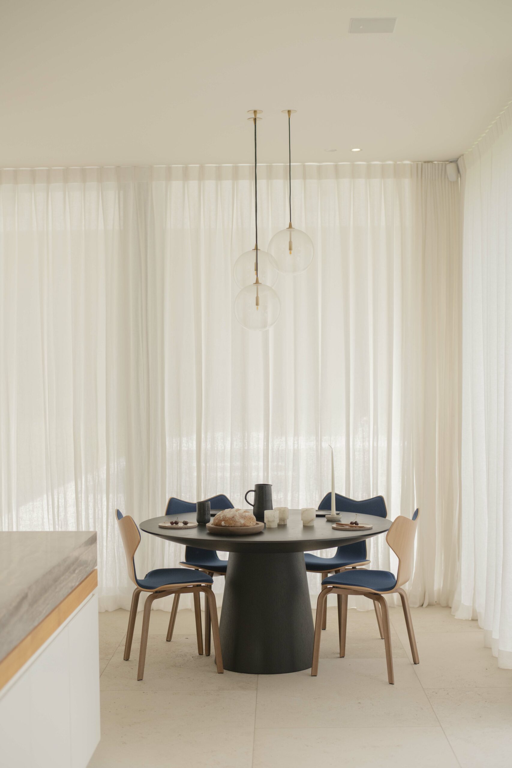 Een elegante eethoek met transparante gordijnen, hangende bolvormige lampen en een donkere ronde tafel. Blauwe stoelen en minimalistische decoratie versterken de serene sfeer.