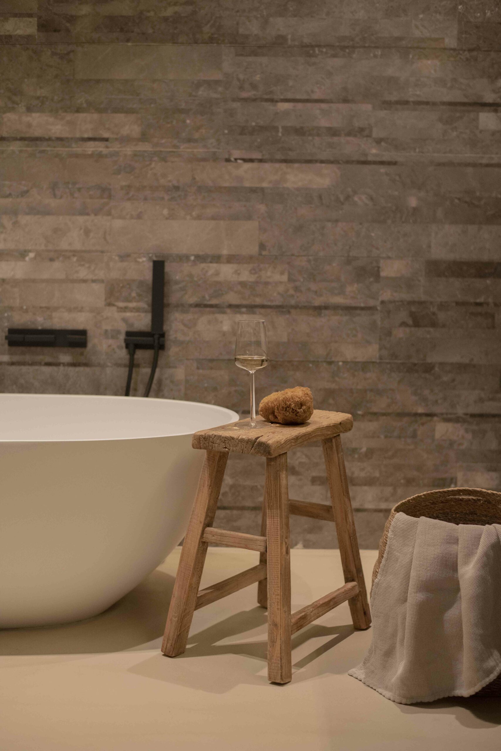 A serene bathroom setting with a stone wall backdrop. A sleek white tub is accompanied by a rustic wooden stool holding a glass of wine and a natural sponge, with a woven basket nearby.