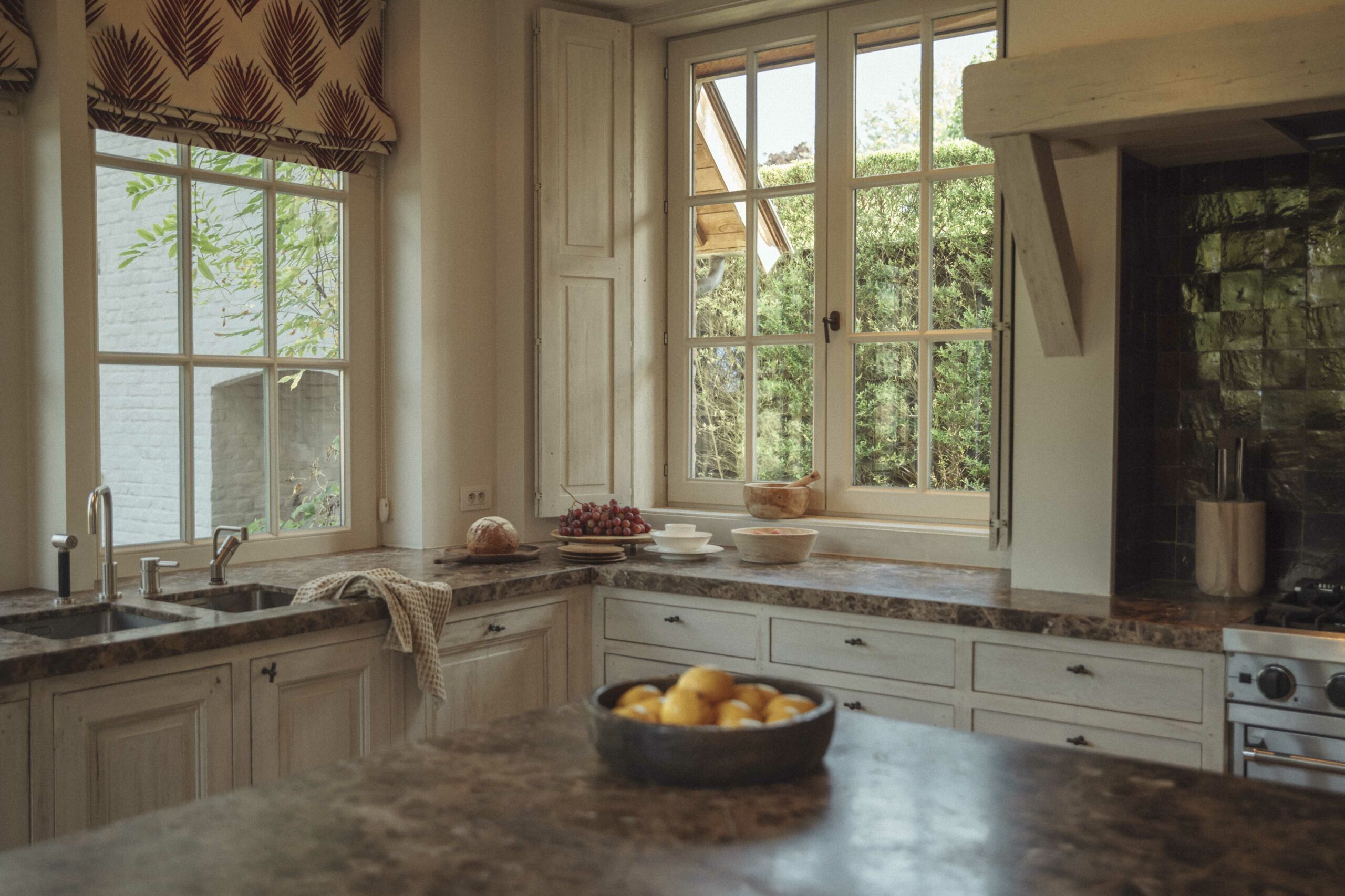 A cozy kitchen with granite counters, rustic white cabinets, modern fixtures, and large windows. Bowls of fruit add a homely touch.
