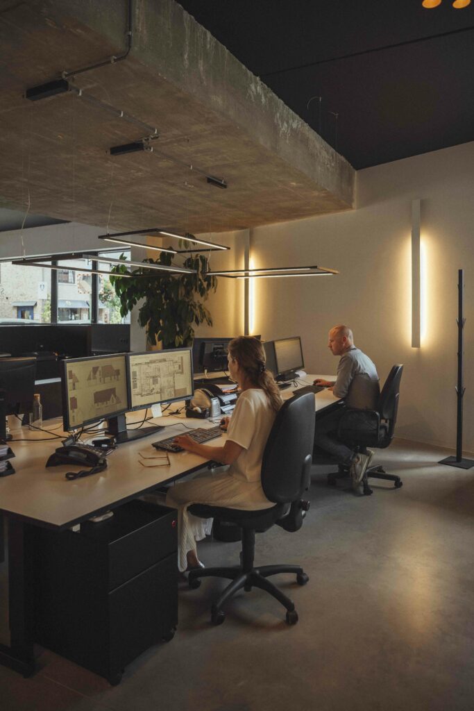 Two professionals working at computer desks in a modern office with exposed concrete ceiling and warm lighting.