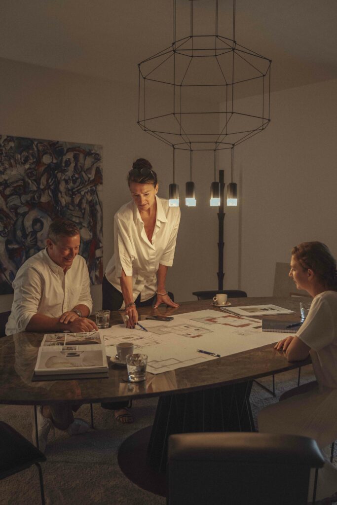 Three individuals gathered around a table reviewing architectural plans in a dimly lit room with a geometric chandelier overhead. A prominent artwork hangs on the wall in the background.