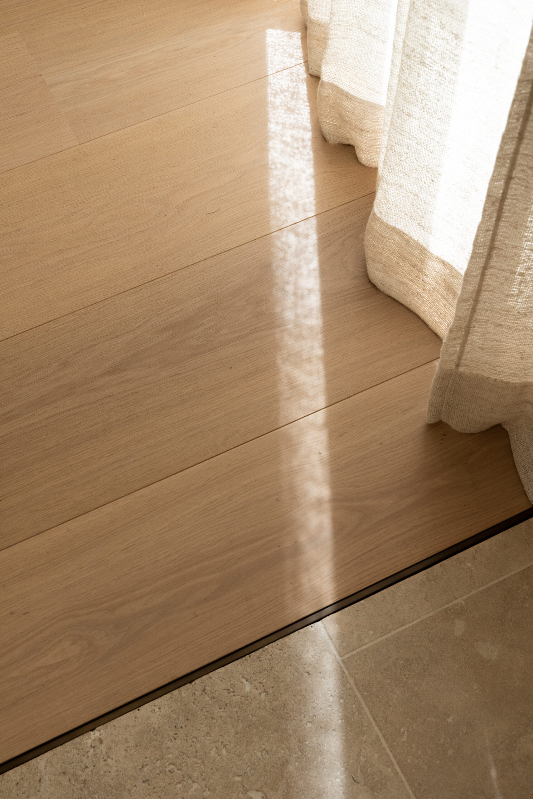 Warm sunlight shines on wooden flooring, meeting a curtain's edge and stone tiles. A serene ambiance prevails.