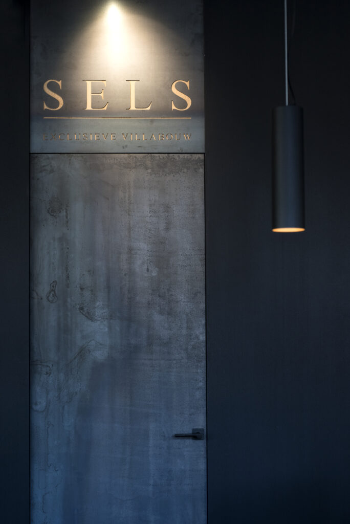 An atmospheric play of light illuminates the "SELS" logo, hinting at exclusive villa construction, while a minimalistic pendant light adds a touch of modern elegance.