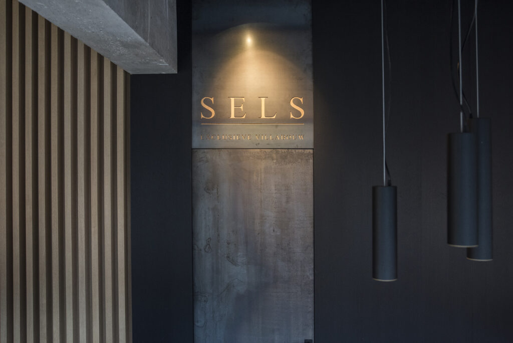 A modern interior with a wooden slatted wall on the left. There's a sign on a concrete-like surface reading "SELS" above "EXCLUSIEVE VILLABOUW".
