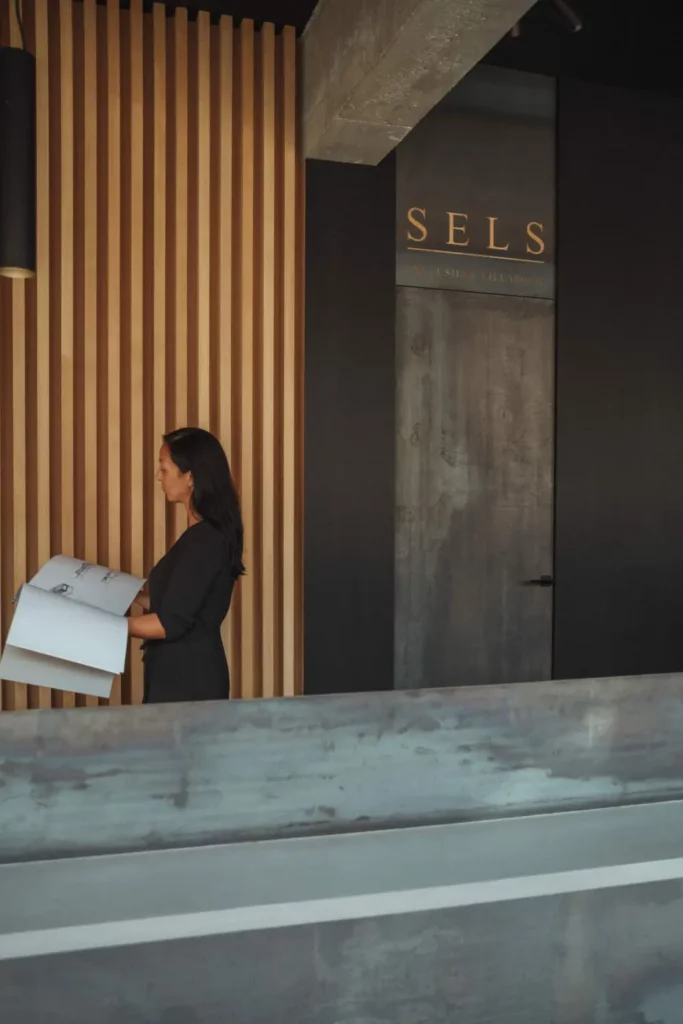 A woman in a black dress holding architectural plans stands beside a striped wooden wall with the word "SELS" on an adjacent dark panel.