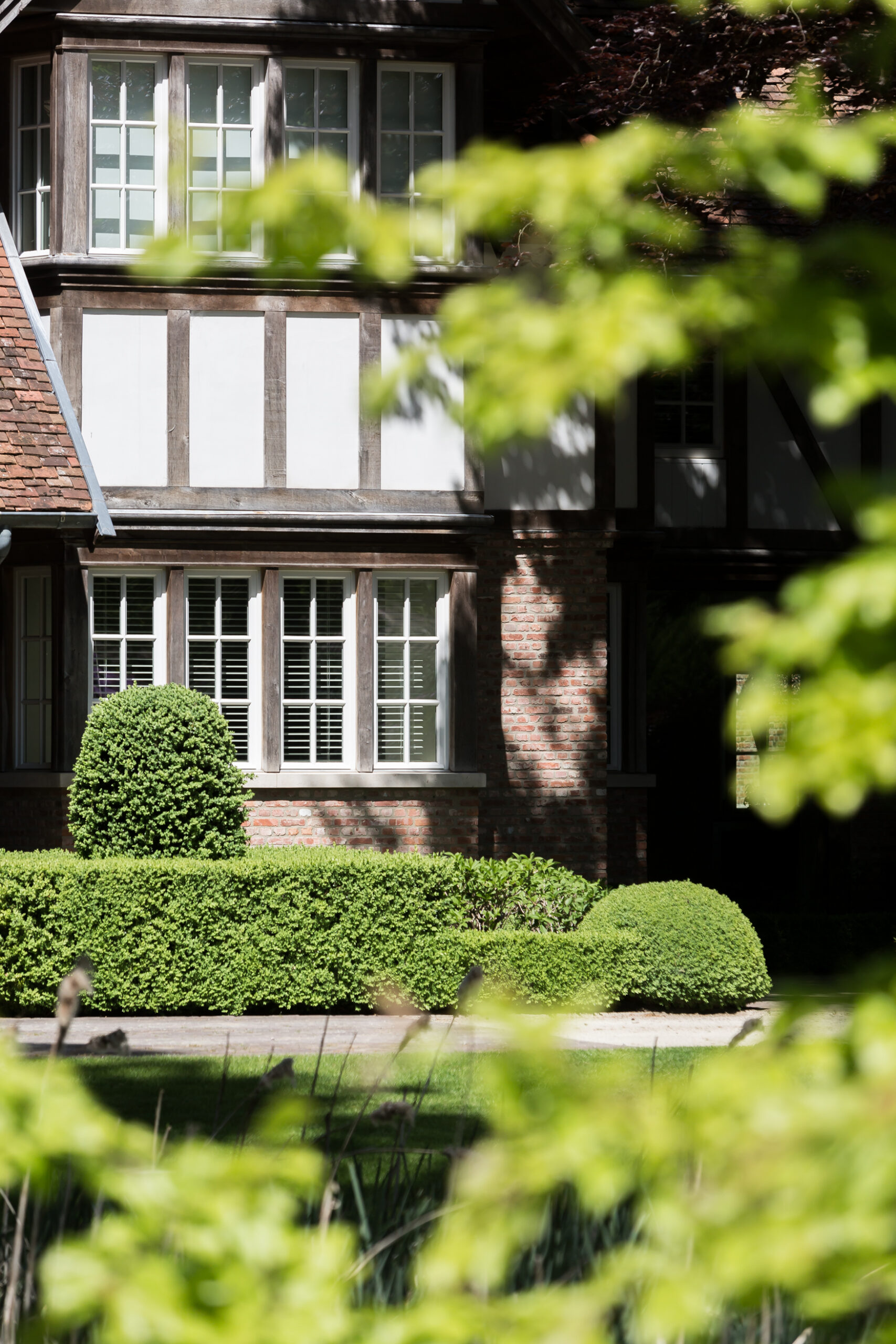 A charming view of a Tudor-style home featuring wooden beams and brickwork. The scene is framed with lush greenery, including manicured bushes in the foreground. The sunlight creates a warm ambiance, casting shadows on parts of the building.