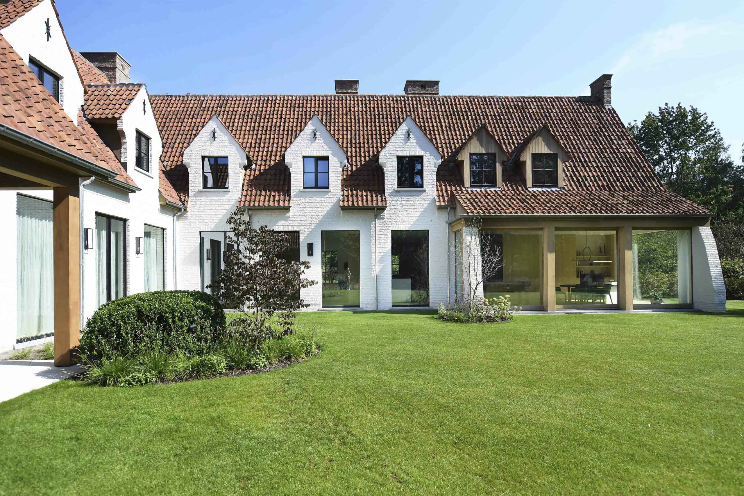 A beautiful white brick house with a tiled roof, featuring a modern glass extension. The well-manicured garden includes a lush green lawn and a variety of plants.