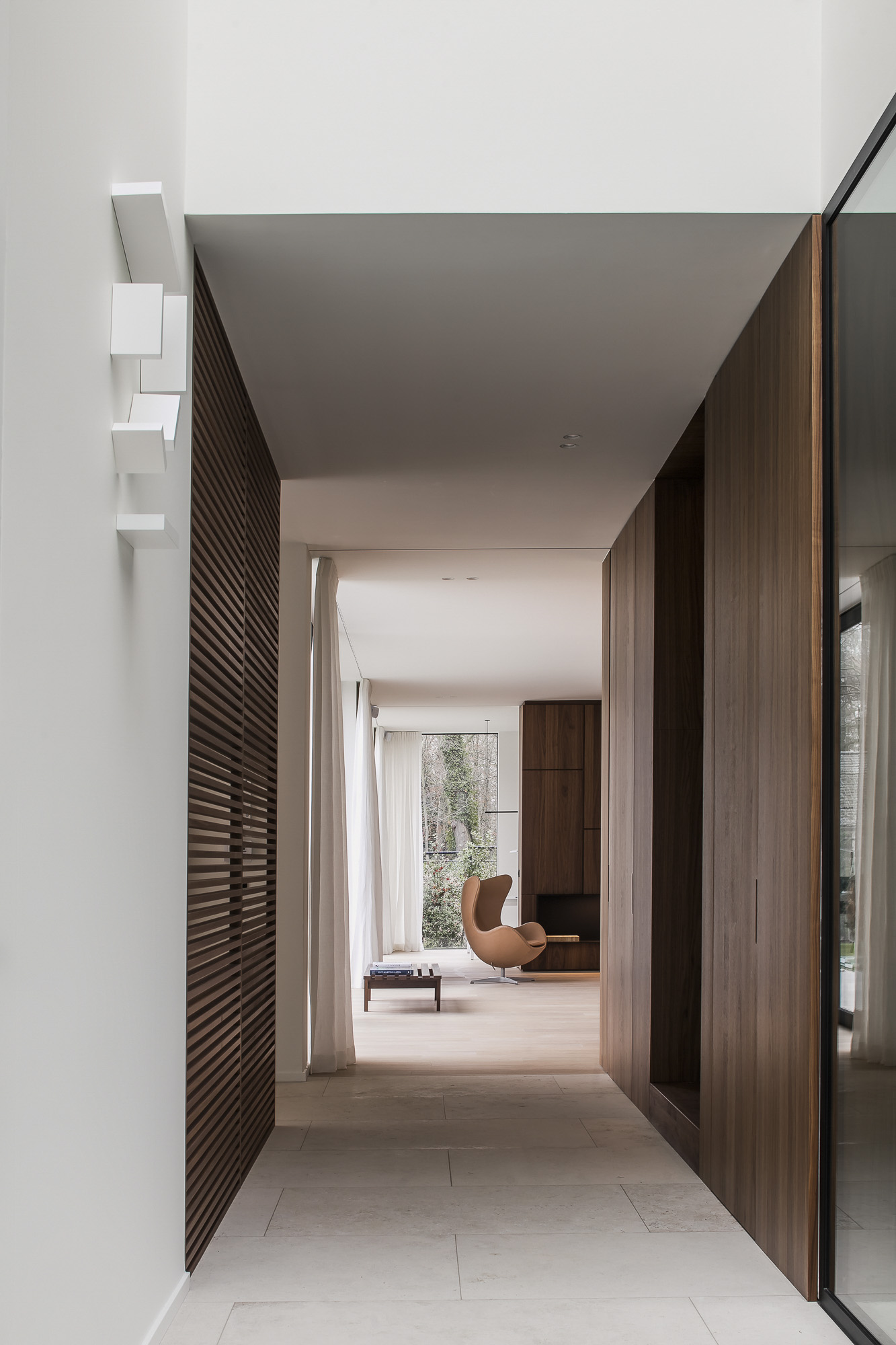 Een moderne binnenruimte met een unieke witte wandlamp, houten accenten, een groot raam en een bruine stoel. Het ontwerp is minimalistisch en verfijnd.