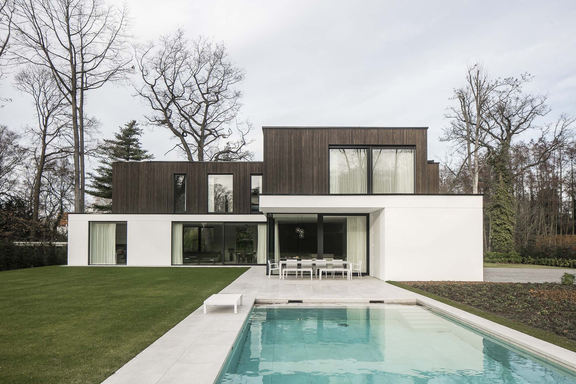 Een modern huis met twee niveaus en gevels van hout en wit. Een smetteloos zwembad op de voorgrond vult het groene gazon aan en hoge bomen omlijsten het terrein.