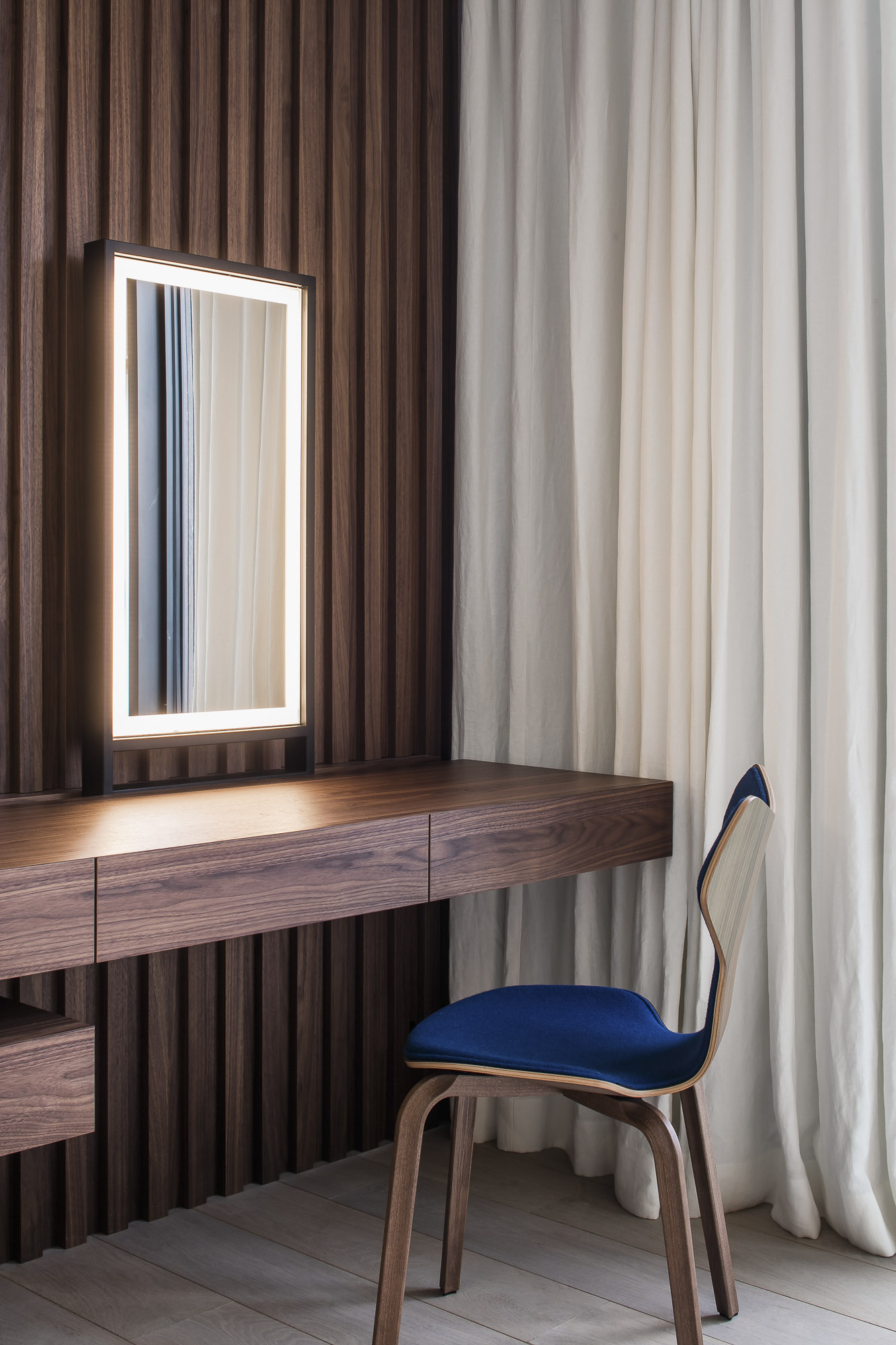 Elegant wooden vanity with an illuminated mirror, paired with a blue-cushioned chair, set against draped white curtains.