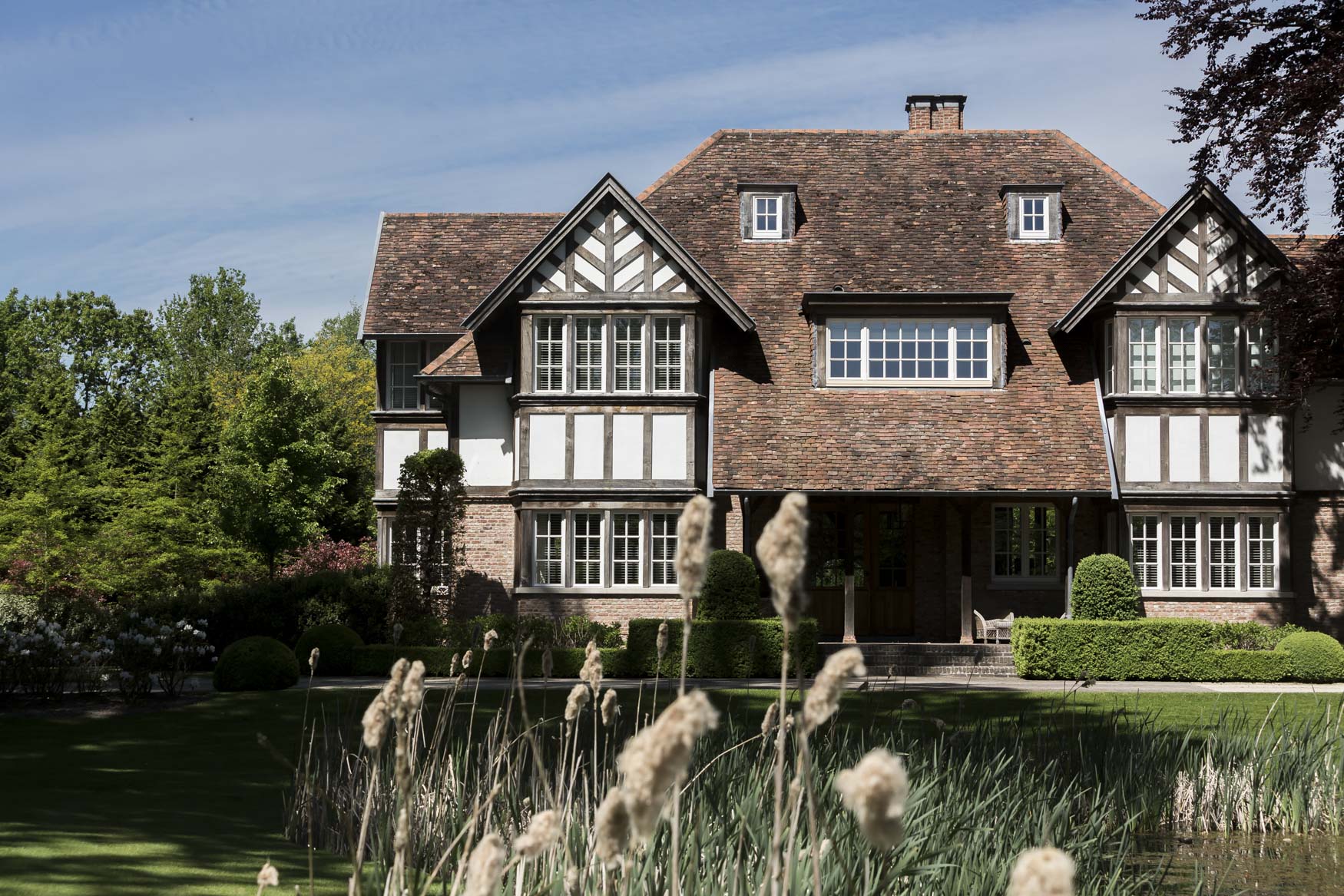 Een huis in Tudorstijl met houten balken en bakstenen muren staat te midden van weelderig groen. Hoge grassen wiegen in de voorgrond, wat diepte toevoegt aan de serene omgeving.