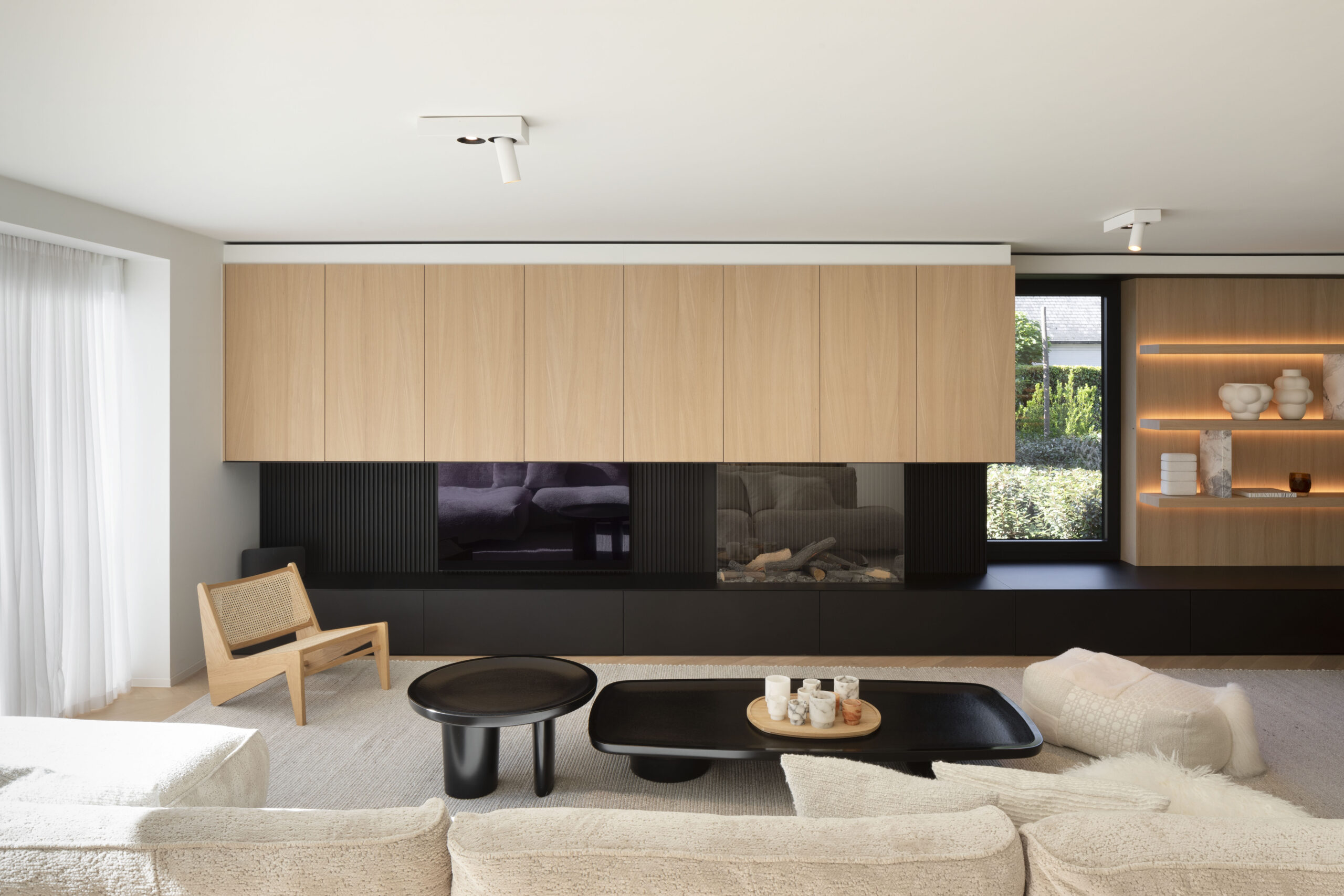 Modern living room with wooden accents, black tables, and lighted shelves.