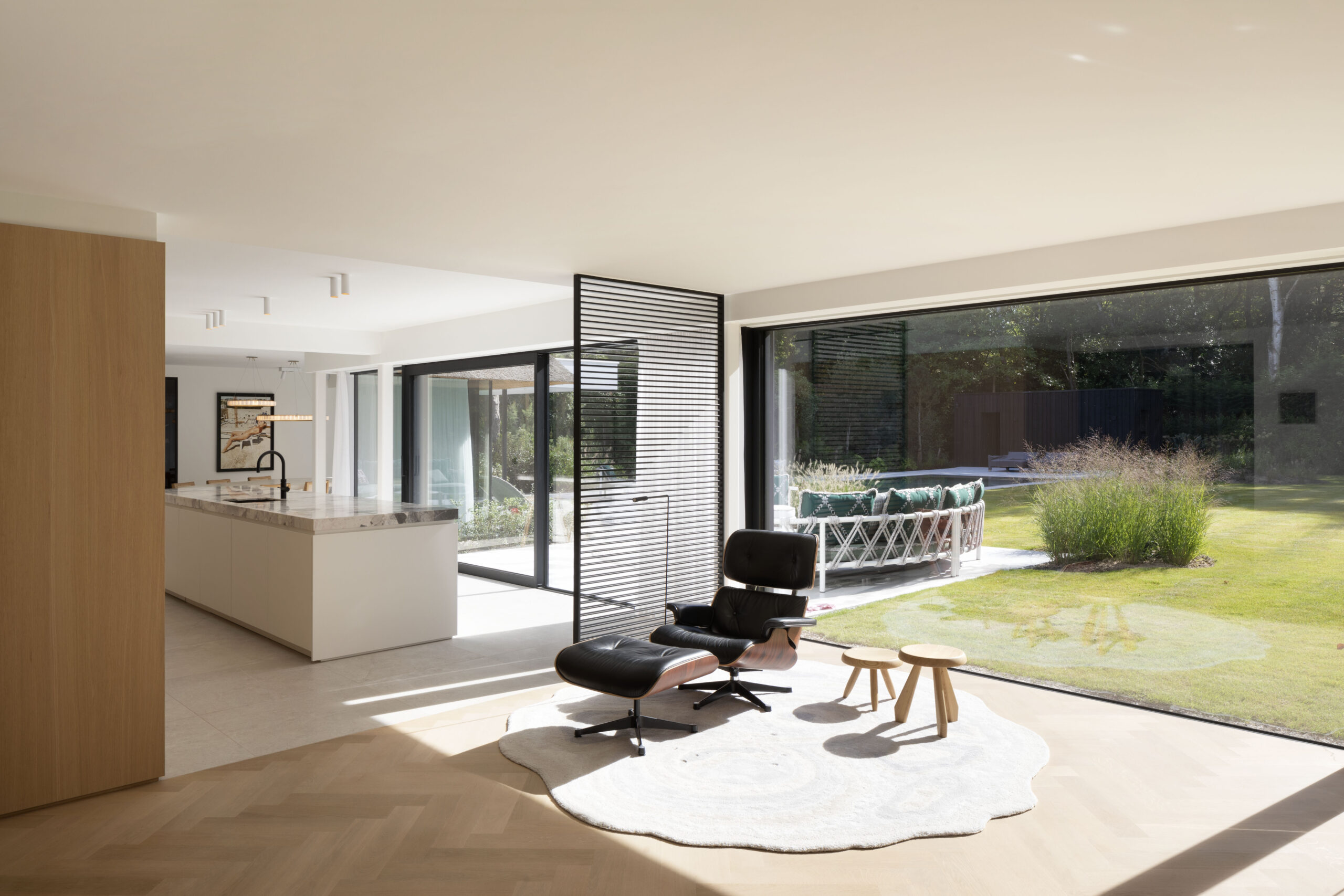Een lichte woonruimte met grote ramen, een moderne zwarte stoel, een wit keukeneiland en lichte houten vloeren.