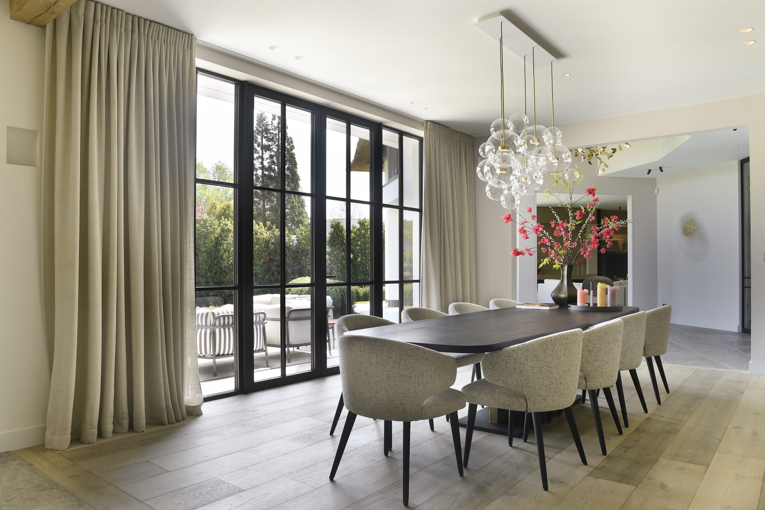 Modern dining room with large windows, elegant chandelier, and a wooden table surrounded by upholstered chairs.