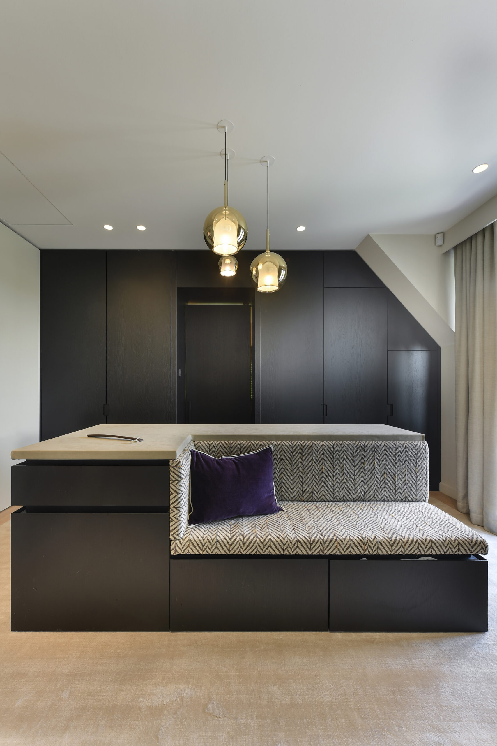 A modern interior space featuring a built-in seating area with a patterned cushion and a purple pillow. Above, there are three pendant lights with glass globes. Adjacent is a dark cabinetry unit, and the floor has a beige finish.