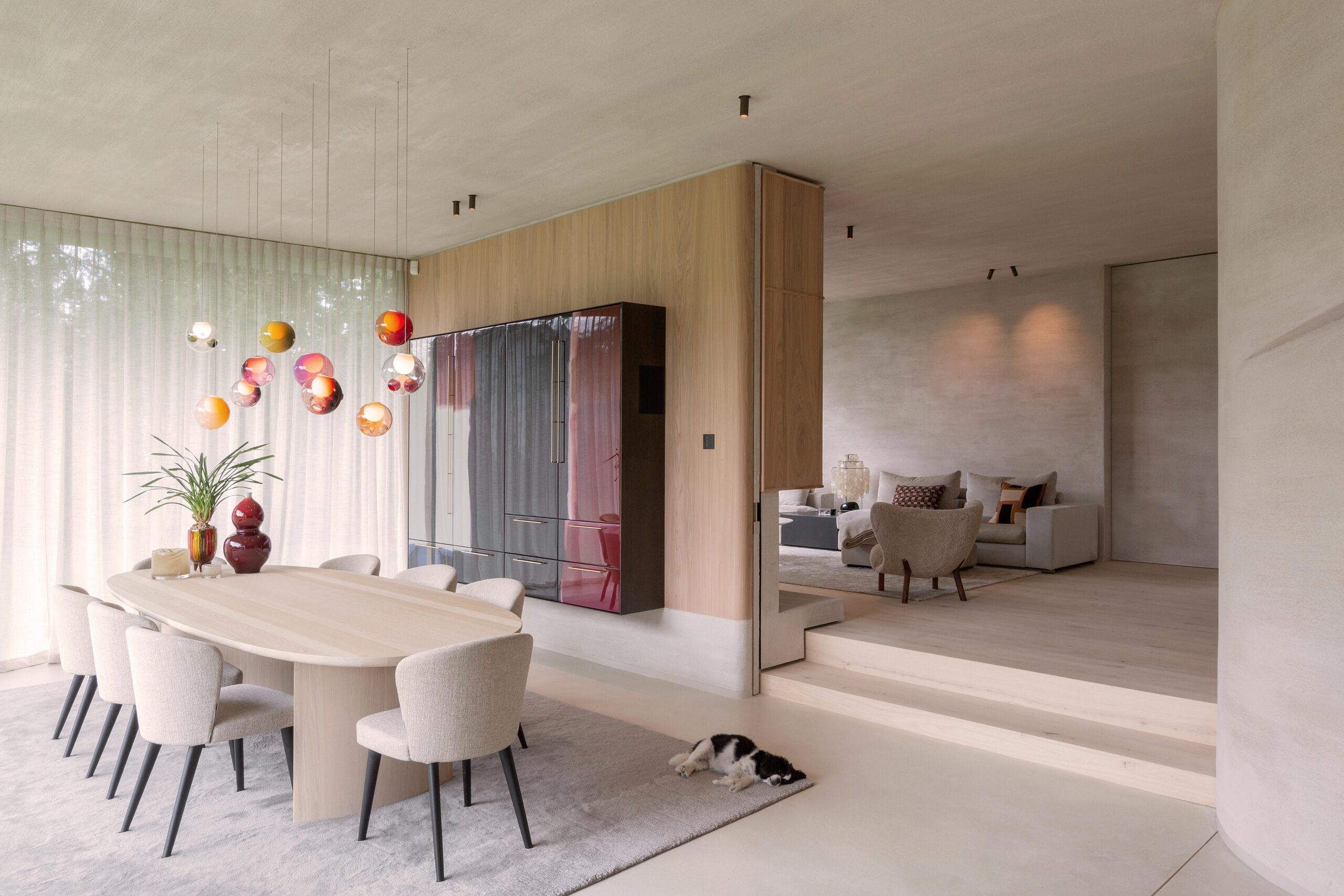 Elegant dining area with a wooden table, upholstered chairs, and colorful suspended glass pendant lights. The room seamlessly connects to a cozy living space with muted tones, and a small dog rests on the rug.