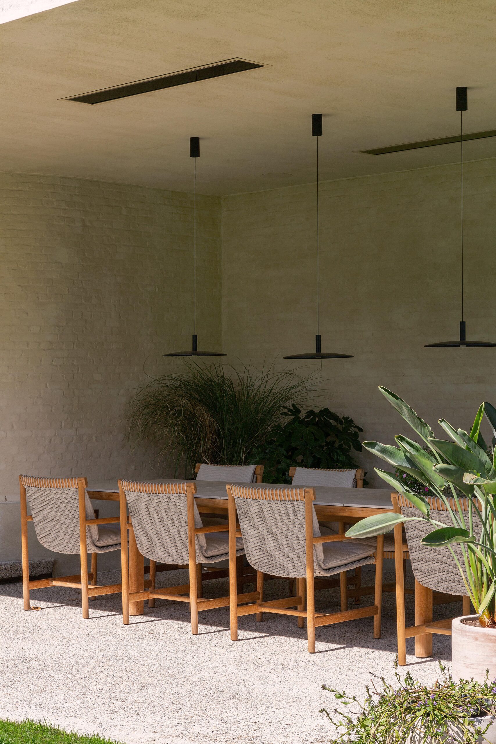 Een buiten eetgedeelte met houten stoelen, moderne hanglampen, een witte bakstenen muur en potten met planten.