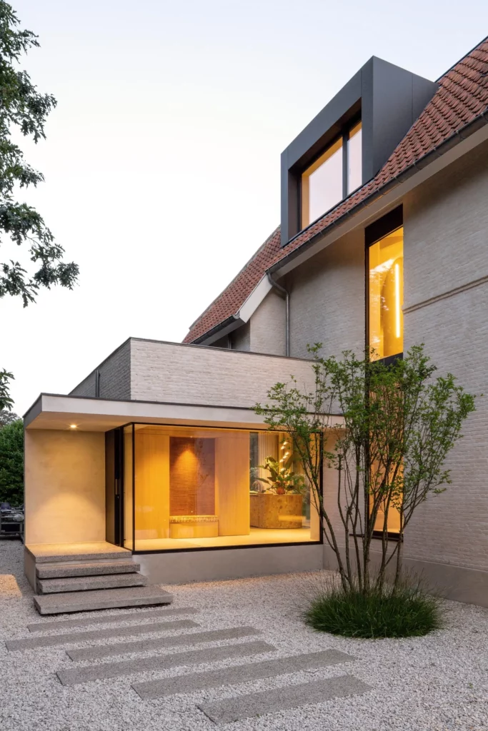 A modern two-story house with a combination of brick and sleek black finishes, illuminated interiors, and a landscaped garden featuring stepping stones and a young tree.