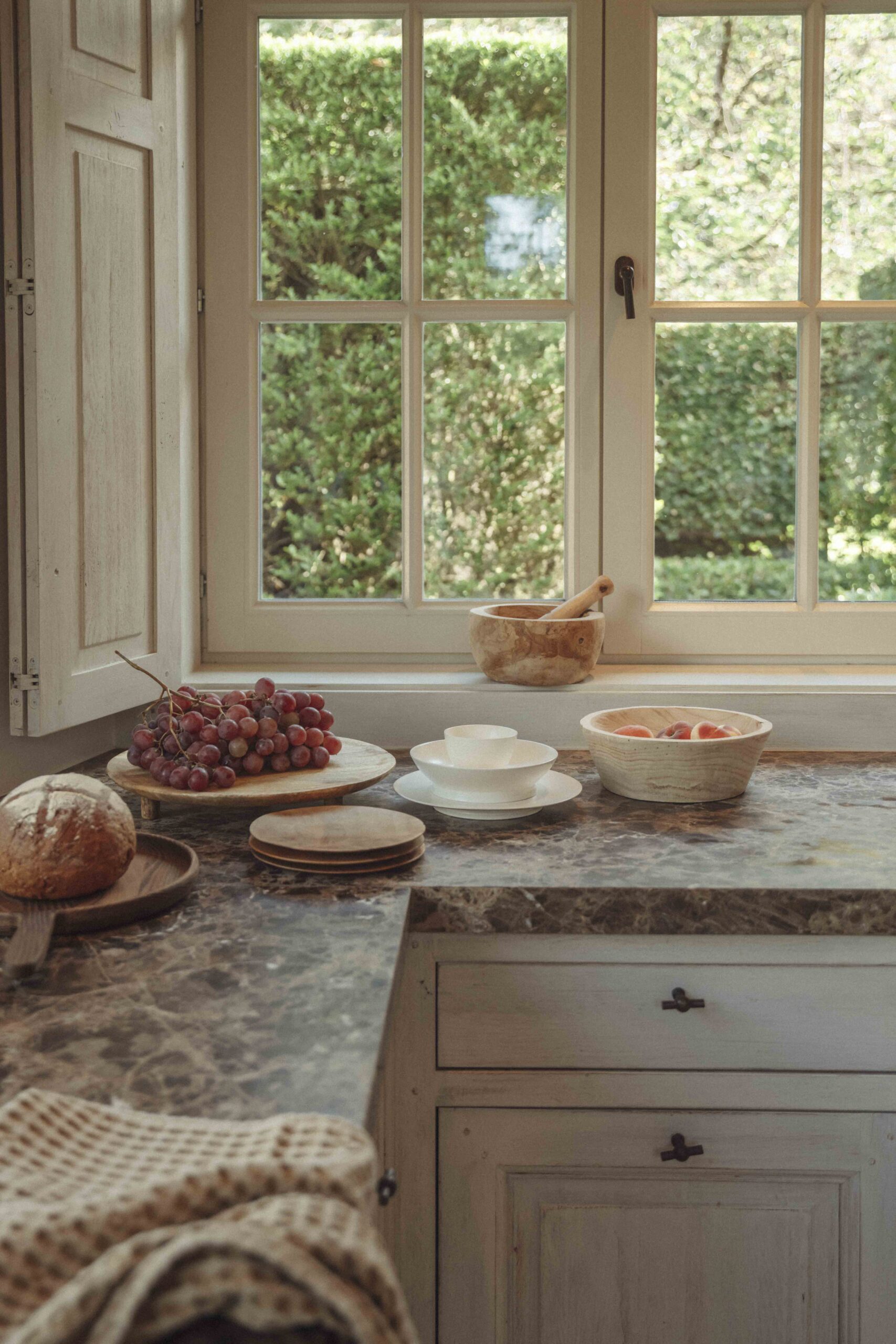 Rustic kitchen with grapes, bread, peaches, and dishes on a marble counter by a window with greenery outside.