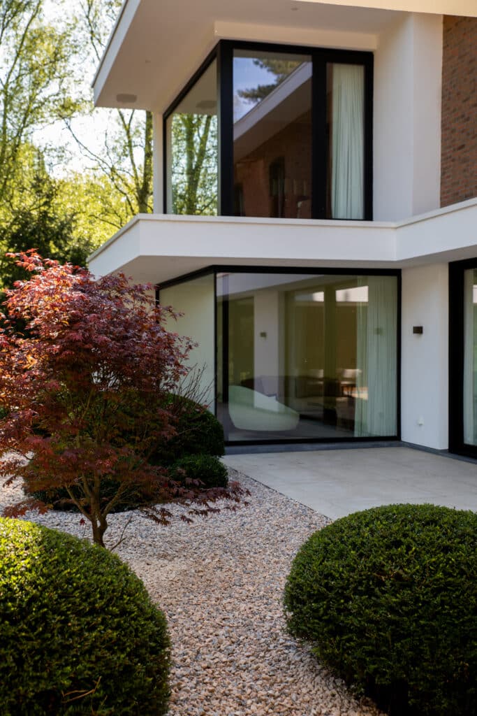 Moderne witte woning met grote glaspartijen, omringd door een verzorgde tuin met struiken, boom en grindpad.