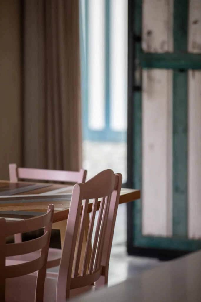 Houten eettafel met stoelen naast een raam met groene en witte luiken in natuurlijk daglicht.