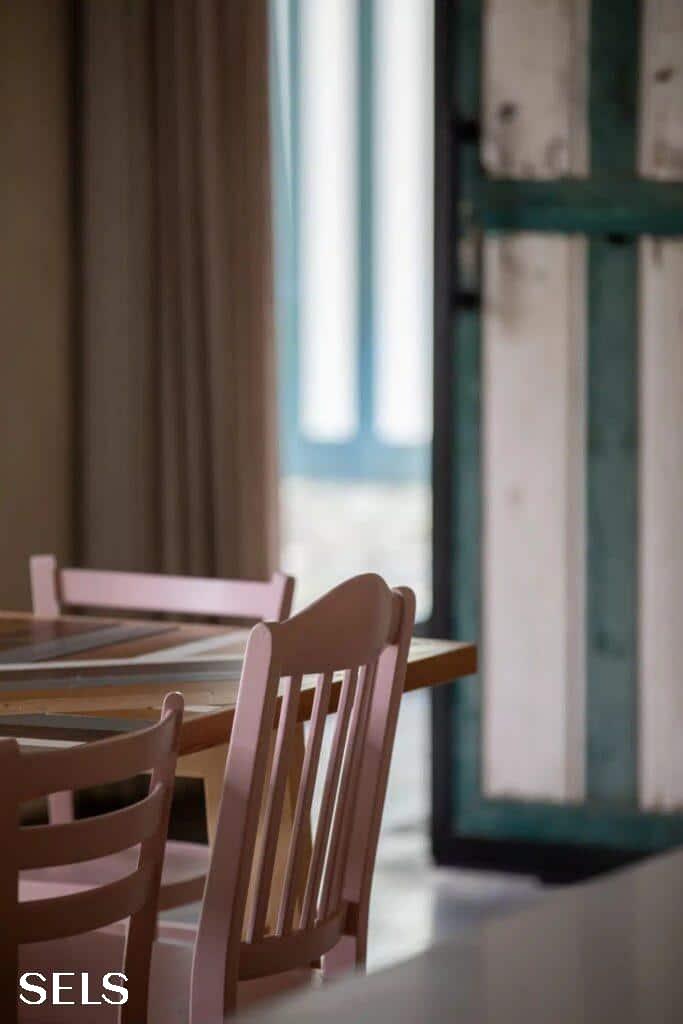Houten eettafel met stoelen naast een raam met groene en witte luiken in natuurlijk daglicht.