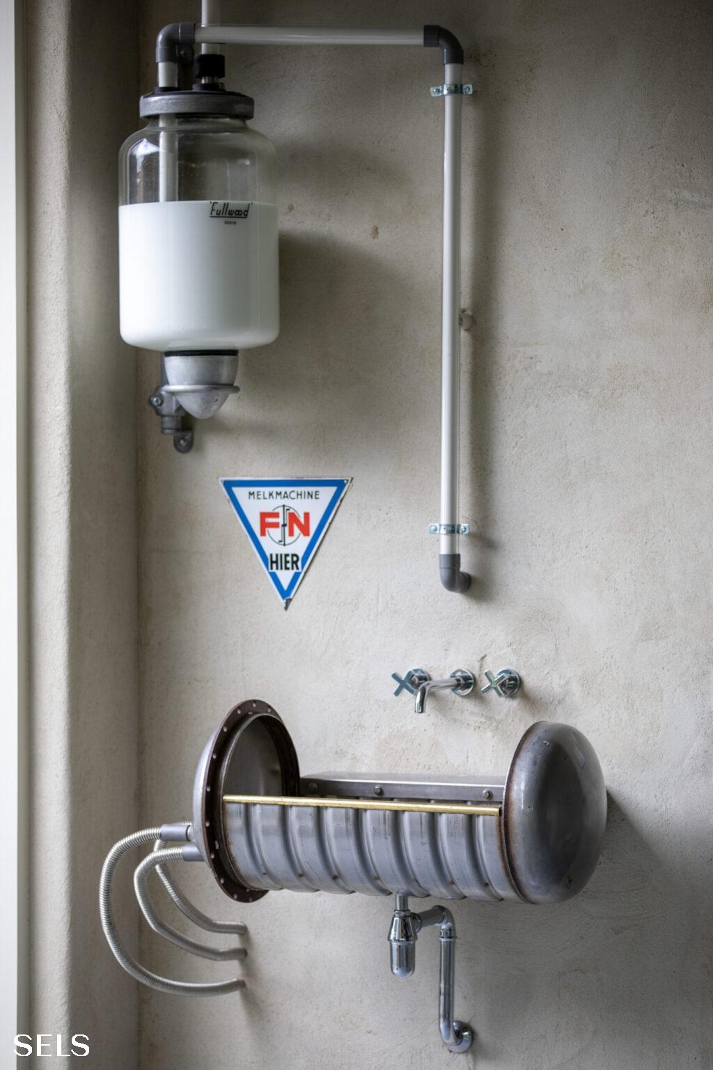 Wandgemonteerde installatie met cilindervormige container bovenaan en horizontale metalen unit met platen eronder, naast een waarschuwingsbord met pijl omlaag.